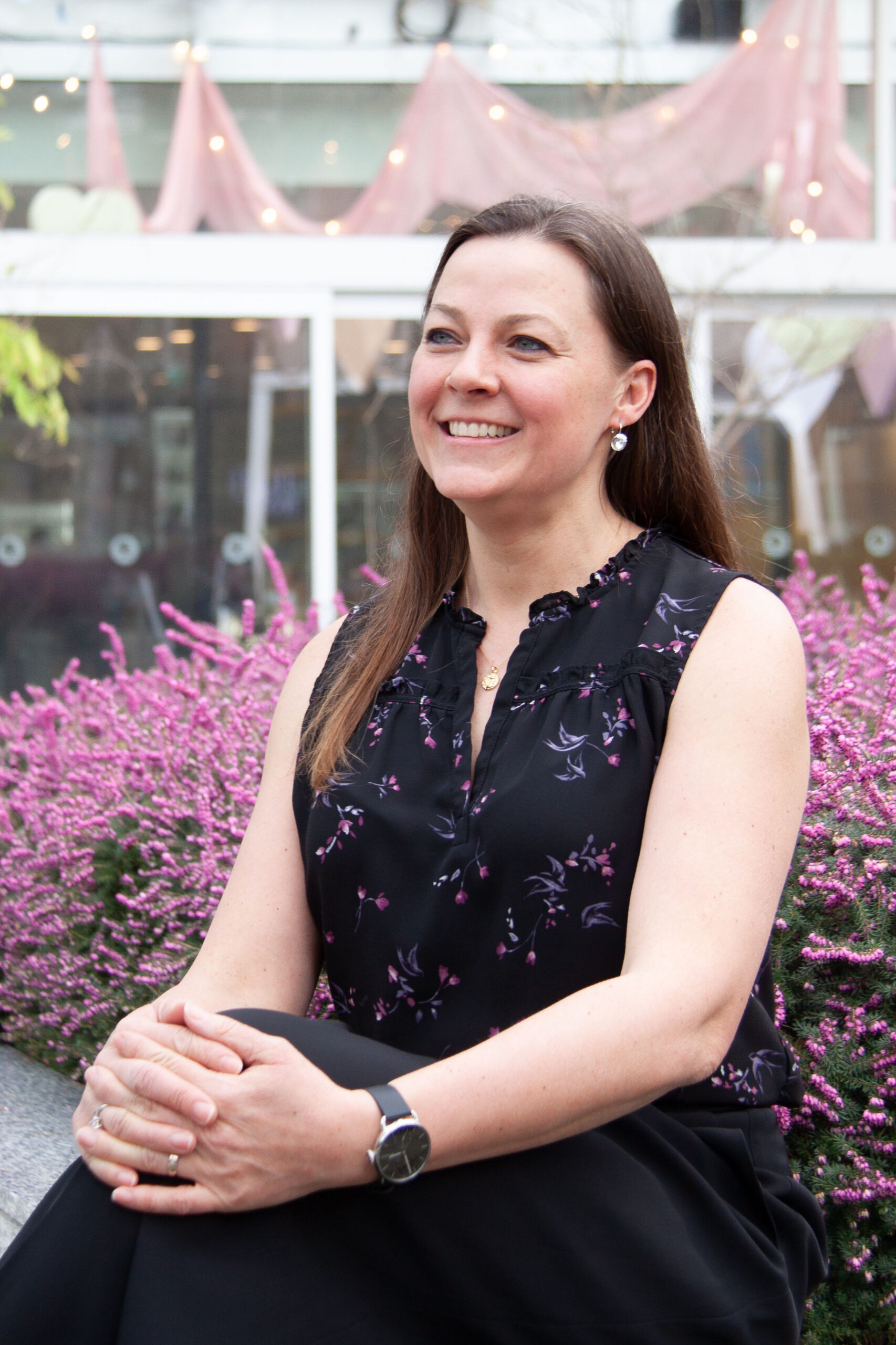 Elegant woman smiling outdoors in garden setting with pink flowers and decorative banners.